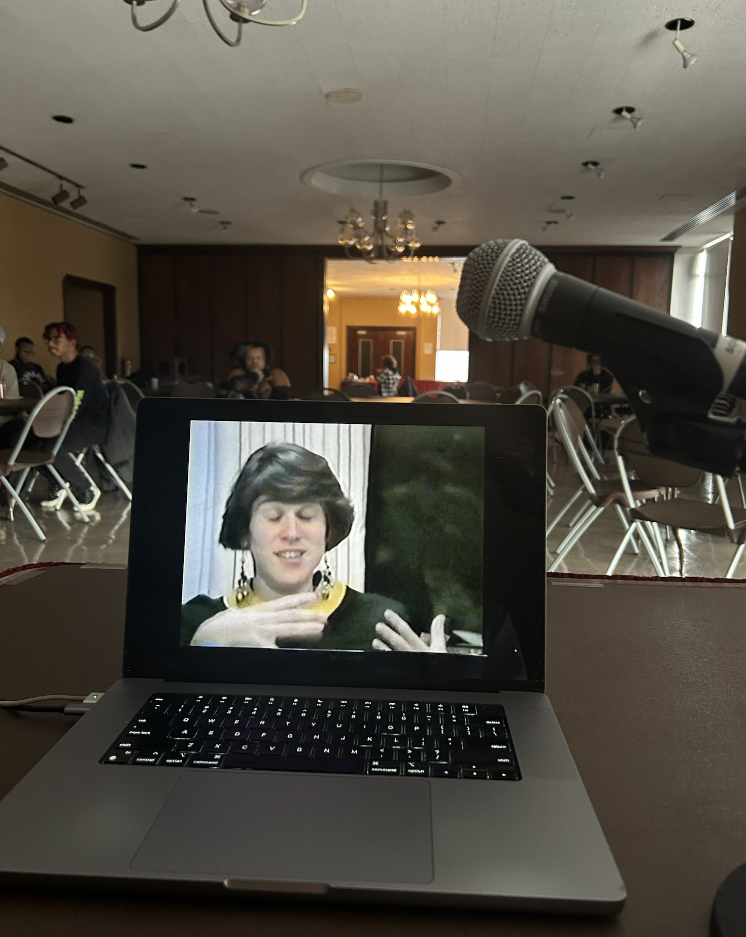 VIdeo screen and microphone in the foreground, audience seen in the distance