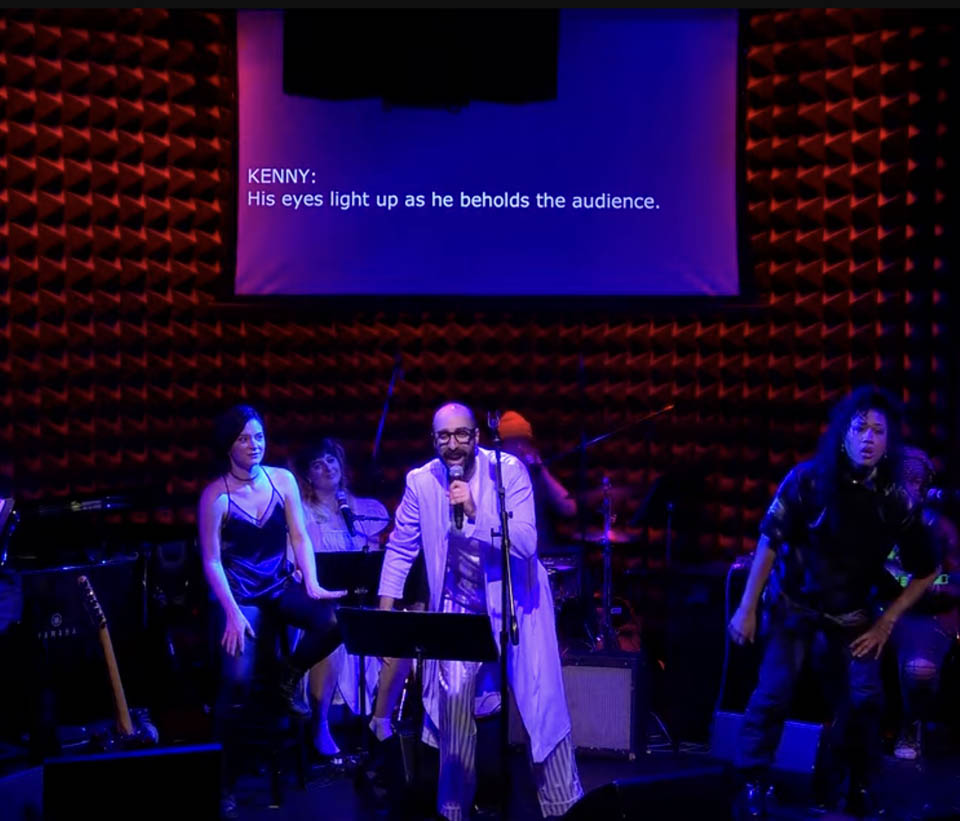 Ron Shalom as Dan with the words, "KENNY: His eyes light up as he beholds the audience." on the projection above the stage
